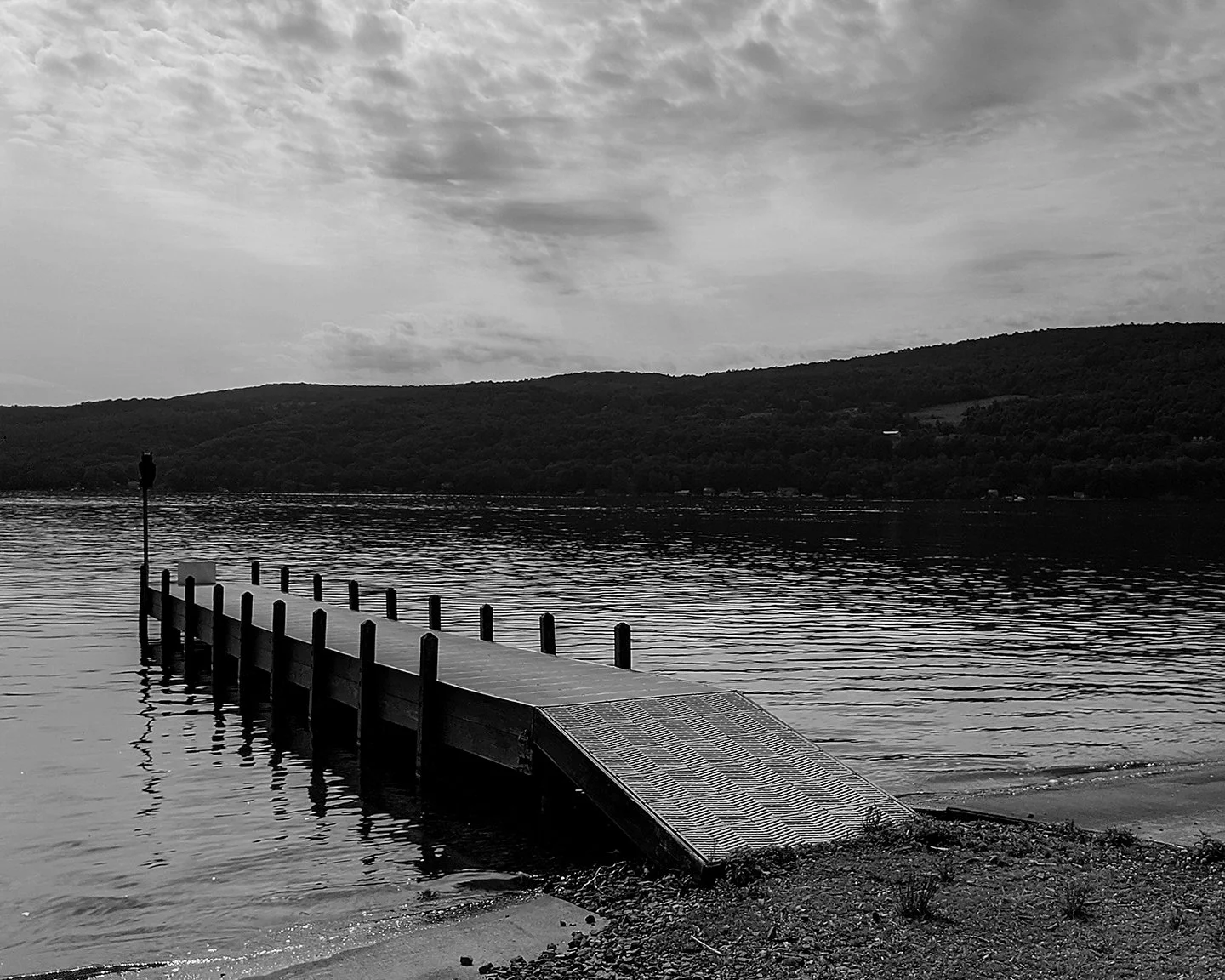 Black-and-white photograph of a dock extending into water beneath wooded hills and a cloudy sky.