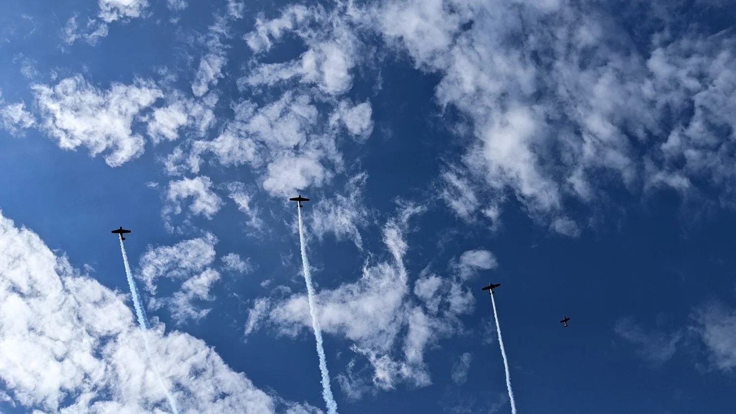 Four aircraft flying in formation through a blue, cloud-filled sky with white smoke trails.